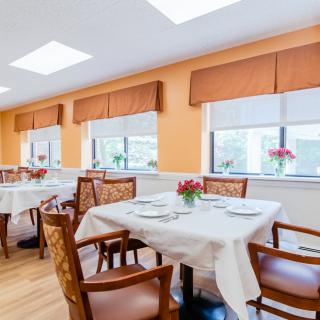 A bright dining room with tables set for a meal, featuring flowers and large windows.