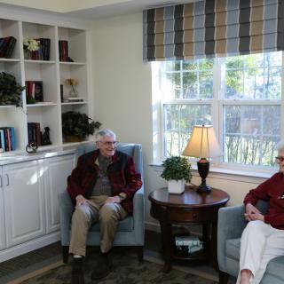 Two elderly individuals sit comfortably in a cozy living room with a bookshelf and window.
