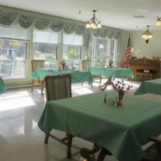 A well-lit dining room with tables covered in green tablecloths, surrounded by chairs, and large windows letting in natural light.