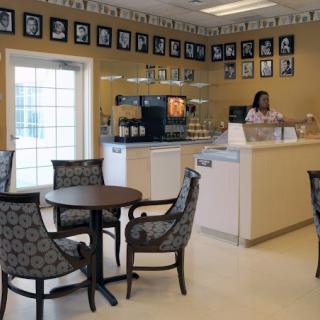 A cozy cafe interior featuring several tables and chairs, a counter with a person standing behind it, and walls adorned with framed photos.