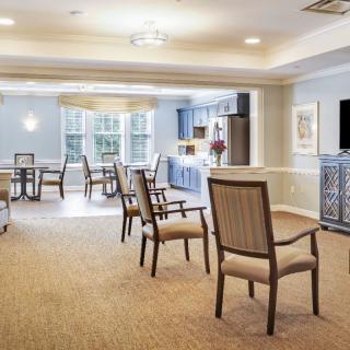 A well-lit dining and kitchen area with chairs and tables arranged on a carpeted floor.