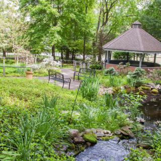 A serene garden featuring a gazebo, benches, and a gentle stream flowing amidst lush greenery.