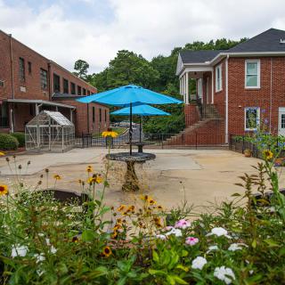 A serene courtyard with brick buildings, colorful flowers, and an umbrella-covered seating area.