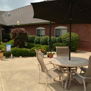 A cozy patio with a table and chairs under an umbrella, surrounded by well-maintained gardens and a brick building in the background.