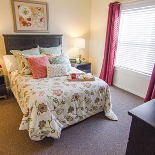 A cozy bedroom with a floral bedspread and decorative pillows, featuring warm lighting and natural light from the window.