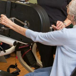 An elderly person engaging in physical exercise using gym equipment, assisted by a trainer.