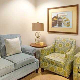 A cozy living room corner featuring a blue armchair, a floral patterned armchair, a side table with a lamp, and wall art.