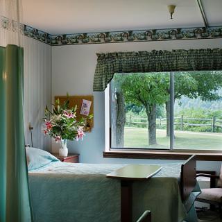 A hospital room with a bed next to a window, offering a view of trees and greenery outside.