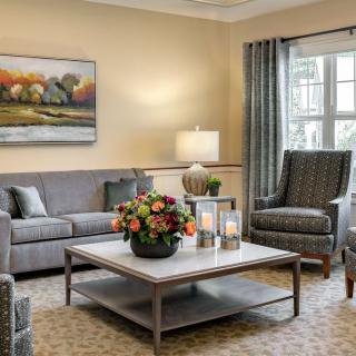 A cozy living room with elegant furniture, featuring a gray sofa, armchairs, and a coffee table adorned with a floral centerpiece. The room is complemented by artwork on the wall, soft lighting from a lamp, and large windows with curtains.