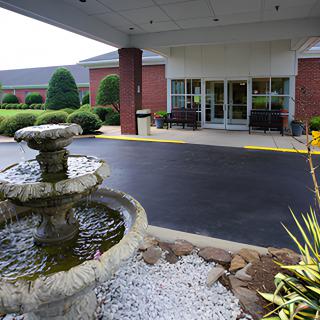A building entrance with a paved driveway, featuring a decorative fountain and well-maintained landscaping.