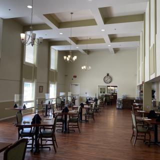 A spacious dining hall with high ceilings, featuring multiple tables and chairs, large windows, chandeliers, and a wooden floor.