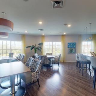 A well-lit dining area with wooden floors, featuring tables and chairs, surrounded by windows and indoor plants.