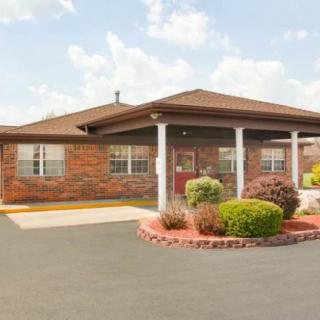 A brick building with a covered entrance, surrounded by neatly landscaped bushes and a parking lot under a cloudy sky.