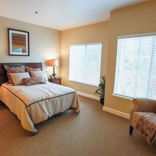 A cozy bedroom with a neatly made bed, two windows, a lamp, and an armchair.