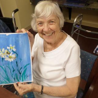 An elderly woman smiling as she holds a painting of flowers.