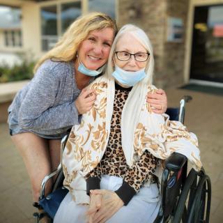 A smiling elderly woman in a wheelchair is accompanied by another smiling person outdoors.