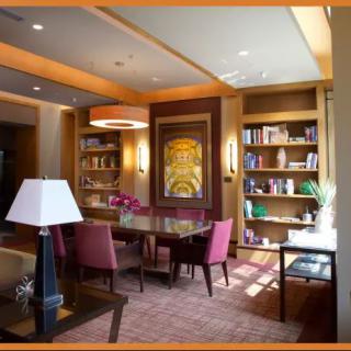A cozy library room featuring a large table surrounded by chairs, with bookshelves and decorative lighting.