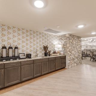 A modern kitchen with a countertop featuring coffee dispensers, leading to a dining area with a stone wall accent.