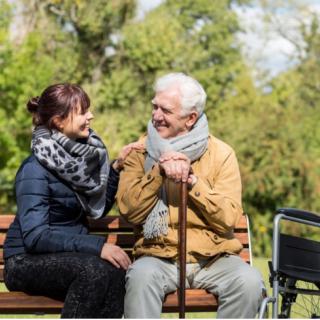 An elderly man with a cane and a younger woman sit on a park bench having a conversation, with a wheelchair nearby.