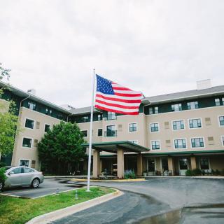 An image of a building with an American flag flying in front, surrounded by a parking lot and trees under a cloudy sky.
