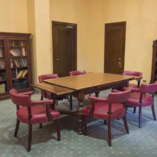 A conference room with a wooden table, surrounded by red chairs, and bookshelves against the wall.