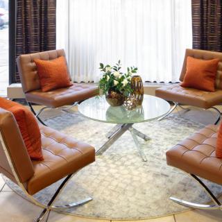 A modern seating area with four brown chairs and a glass table, adorned with decorative plants and orange cushions.