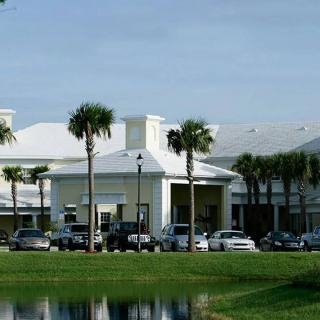A modern building surrounded by palm trees with cars parked in front, reflected in a nearby pond.