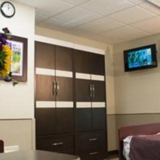 A hospital room featuring a wardrobe, wall-mounted television, bed, and clock.