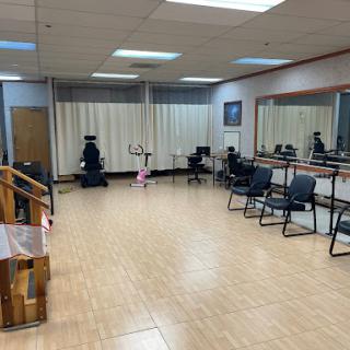 An empty dance studio featuring mirrors, wooden flooring, and chairs arranged around the space.