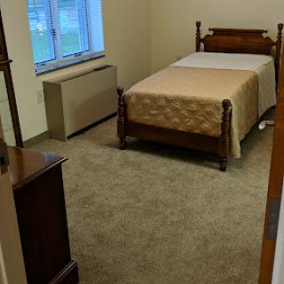 A simple and tidy bedroom featuring a wooden bed, carpeted floor, and a window allowing natural light.