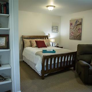 A cozy bedroom with a wooden bed, artwork on the walls, a comfortable armchair, and built-in shelves filled with books.