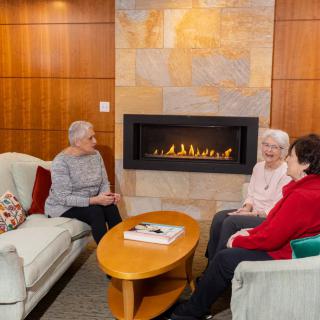 Three seniors enjoy a cozy conversation by the fireplace in a living room setting.