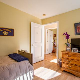 A cozy bedroom featuring a bed, dresser, and floral decorations with warm sunlight streaming in.