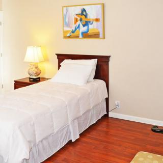 A cozy bedroom features a single bed with white linens, a wooden nightstand with a lamp, and artwork on the wall.