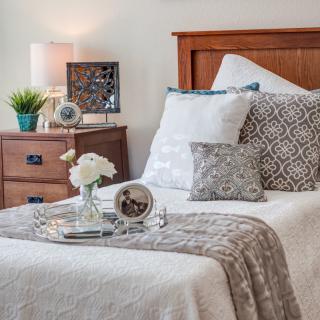 A cozy bedroom with a neatly arranged bed, decorative pillows, and a bedside table featuring a lamp and flowers.