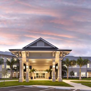 A well-lit entrance of a modern building at dusk with palm trees lining the pathway.