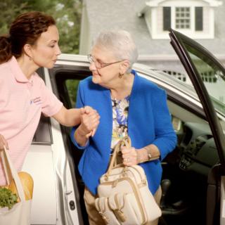 A caregiver assists an elderly woman as she exits a car.