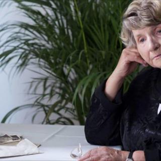 An elderly woman sitting at a table with papers, appearing thoughtful.