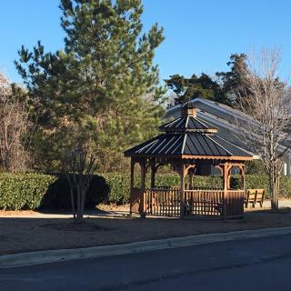 A wooden gazebo in a park surrounded by trees and greenery under a clear blue sky.