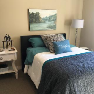 A cozy bedroom with a double bed, featuring decorative pillows and a blue and white color scheme. A painting hangs above the bed, complemented by a nightstand and lamp.