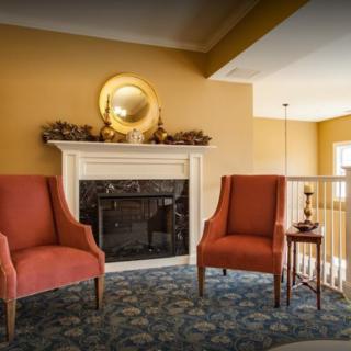 A cozy living room with two red chairs flanking a decorative fireplace, adorned with a round mirror and autumn accents.