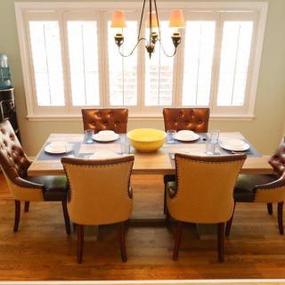 A neatly set dining table with six chairs, illuminated by a chandelier, in a room with large windows and wooden flooring.
