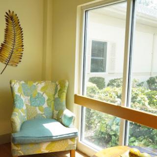 A cozy corner featuring a floral armchair illuminated by natural light, adjacent to a large window with views of greenery.