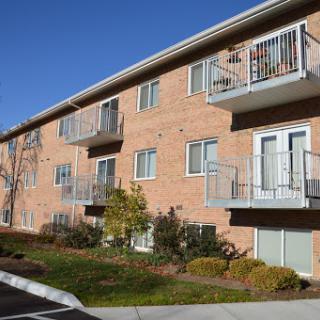 A serene view of a residential apartment building featuring brick construction and balconies, surrounded by well-maintained landscaping under a clear blue sky.