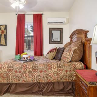 A cozy bedroom featuring a patterned bedspread, wooden furniture, and red curtains, with an air conditioner mounted above the window.