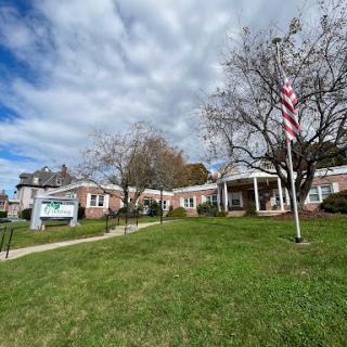 A peaceful scene featuring a building with an American flag in front, surrounded by green grass and a partly cloudy sky.