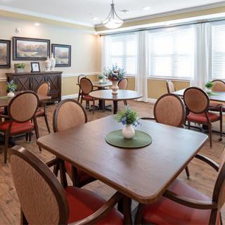 A cozy dining room with neatly arranged tables and chairs, adorned with decorative plants and brightened by natural light from large windows.