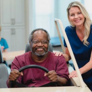 An elderly man smiling while a healthcare worker assists him in a wheelchair simulation.