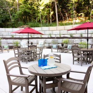 Outdoor patio with tables, chairs, and red umbrellas set against a lush landscaped backdrop.