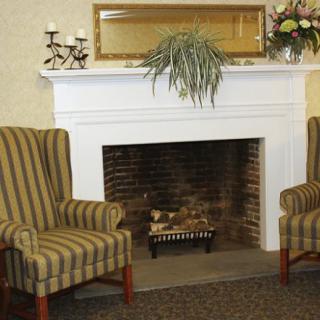 A cozy living room setup featuring a fireplace with a white mantel, flanked by two striped armchairs. Decorative elements include candles and plants.